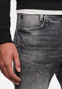 Tmavě šedé džíny z denimu s vybledlým vzhledem a odřenými detaily. Obsahují zapínání na knoflík, kapsy a viditelné švy.