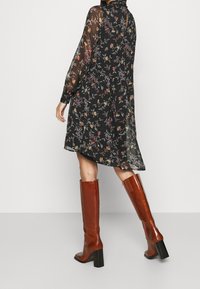 Robe noire à motif floral avec manches longues en voile et col montant, associée à de grandes bottes à talons en cuir marron, mettant en valeur une silhouette fluide.