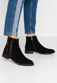 Bottes cheville en daim noir avec une fermeture éclair latérale et un accent en imprimé léopard. Design à talon bas avec une semelle en caoutchouc lisse.
