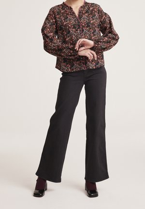 Femme portant un chemisier à manches longues fleuri rentré dans un pantalon noir taille haute à jambes larges et des chaussures noires à talons avec des chaussettes foncées.