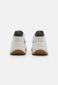 White sneakers with a textured brown heel, featuring a black accent with a logo. The sole has a wavy pattern for traction.