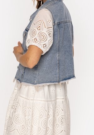 Vrouw die een blauw denimvest draagt over een witte jurk met ingewikkelde oogjeskantpatronen en pofmouwen, gezien van de zijkant en achterkant.