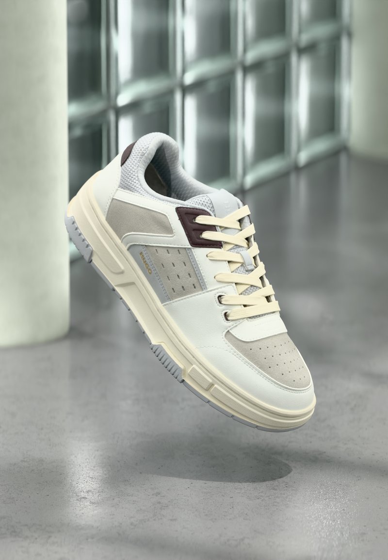 Λευκό και μπεζ αθλητικό παπούτσι με κρεμ κορδόνια, καφέ λεπτομέρειες και διάτρητο σχέδιο στη μύτη, εμφανιζόμενο στον αέρα πάνω από γκρίζο πάτωμα και γυάλινο τοίχο.