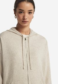 Beige zip-up hoodie i mjukt tyg, med dragsko i huvan, fickor fram och en avslappnad passform. Minimalistisk stil, utan mönster.