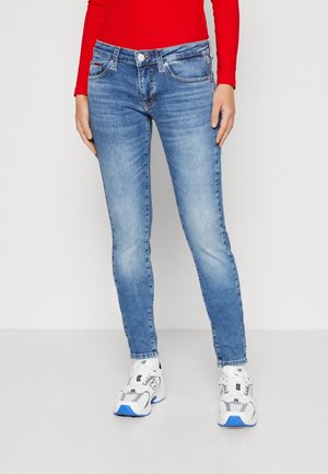 Vaqueros ajustados de color azul claro con un diseño clásico de cinco bolsillos, botón frontal y cierre de cremallera; combinados con zapatillas blancas y una blusa roja.