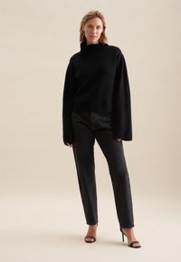 Czarny oversize sweter z wysokim dekoltem, zestawiony z czarnymi prostymi spodniami i czarnymi sandałami na obcasie. Gładka tekstura, nowoczesny design.