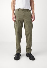 Pantaloni cargo verde oliva realizzati in cotone, con diverse tasche laterali, design a gamba dritta e cuciture rinforzate. Indossati con stivali neri.