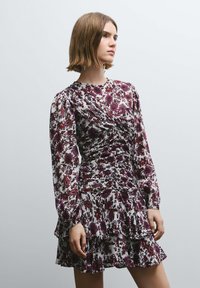 Vestido floral de manga larga, con base blanca, diseños en púrpura y rojo, detalles fruncidos y un dobladillo con volantes.