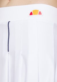 Prenda blanca plisada con un cordón azul marino. Presenta un logo redondo en naranja y rojo en la cintura. Tejido liso y ligero.