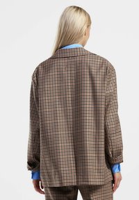 Giacca a quadri marrone e beige con una vestibilità rilassata, spalle larghe e polsini di colore blu. La vista posteriore mette in evidenza il tessuto strutturato.