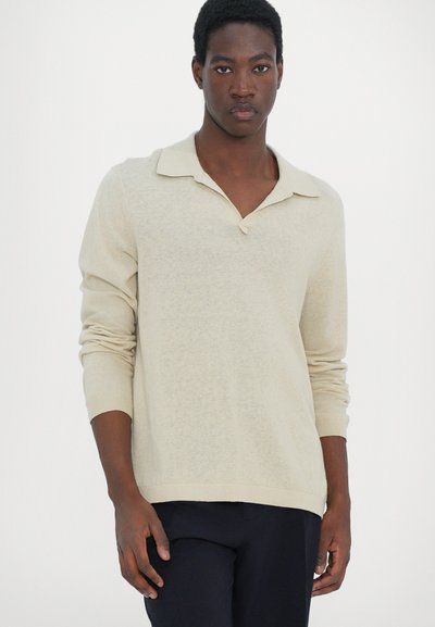 Beige langermet polo med avslappet krage og knappeakcent. Myk tekstur og tettsittende mansjetter. Brukt over mørke bukser.