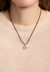 Gros plan d'une personne portant un collier de perles avec un pendentif circulaire en or orné de petites pierres précieuses, sur un pull beige clair.