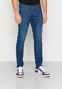 Jeans de ganga ajustados azul com uma textura suave, costuras contrastantes e bolsos frontais, combinados com sapatilhas brancas e roxas.