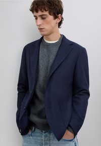 Blazer azul marino oscuro con un acabado texturizado, de dos botones, superpuesto a un suéter de punto gris, combinado con jeans azul claro.