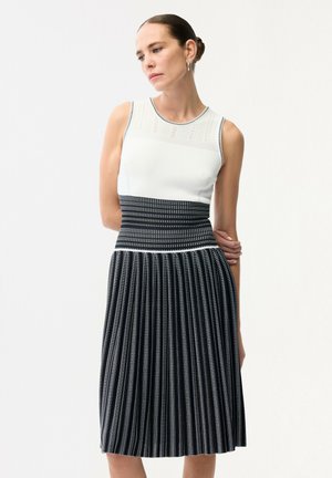 Kvinde iført hvid ærmeløs top og sorte-og-hvide stribede plisserede nederdel med høj talje, håret sat op i knold, kigger ned med armene krydset bag ryggen.