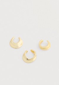 Tres brillantes anillos abiertos de estilo brazalete en oro con superficies suaves y curvadas, dispuestos sobre un fondo gris claro.