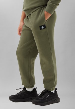 Osoba nosząca oliwkowe dresy Calvin Klein z rękami w kieszeniach oraz czarne trampki Calvin Klein z białymi skarpetkami.
