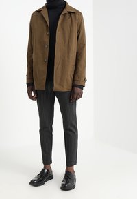 Brun, lätt jacka med notchkrafter och knappstängning, ihop med svart stickad polotröja och randiga grå byxor, avslutad med svarta skor.