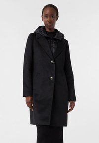 Cappotto nero con finitura texturizzata, dotato di un colletto a ribalta, due bottoni, tasche laterali e un cappuccio imbottito nero attaccato.