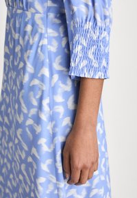 Rochie albastră cu un model abstract alb, având mâneci pliseu și o textură moale și netedă. Se vede o parte a unei mâini odihnindu-se pe material.