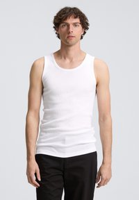 Débardeur en coton blanc avec une texture côtelée et des bretelles larges, porté par une personne aux cheveux courts et bouclés, associé à un pantalon noir.