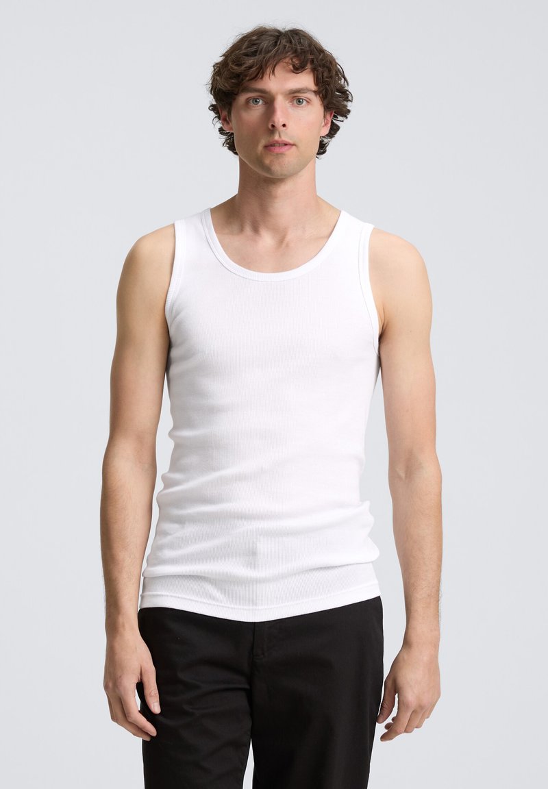 Débardeur en coton blanc avec une texture côtelée et des bretelles larges, porté par une personne aux cheveux courts et bouclés, associé à un pantalon noir.