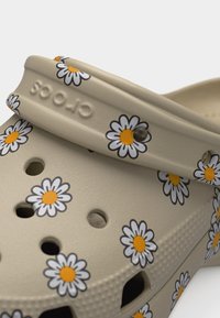Μπεζ παπούτσι Crocs τύπου clog με σχέδια μαργαρίτας και οπές αερισμού, με λουράκι που φέρει ανάγλυφο το λογότυπο Crocs.