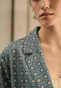 Modrý sako s vzorem, který obsahuje květinový design s černými akcenty. Měkká textilie, klopový límec a viditelné knoflíkové detaily na rameni.