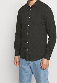 Camicia a maniche lunghe grigio scuro realizzata in tessuto strutturato, con colletto, chiusura frontale con bottoni bianchi. Abbinata a jeans azzurri.