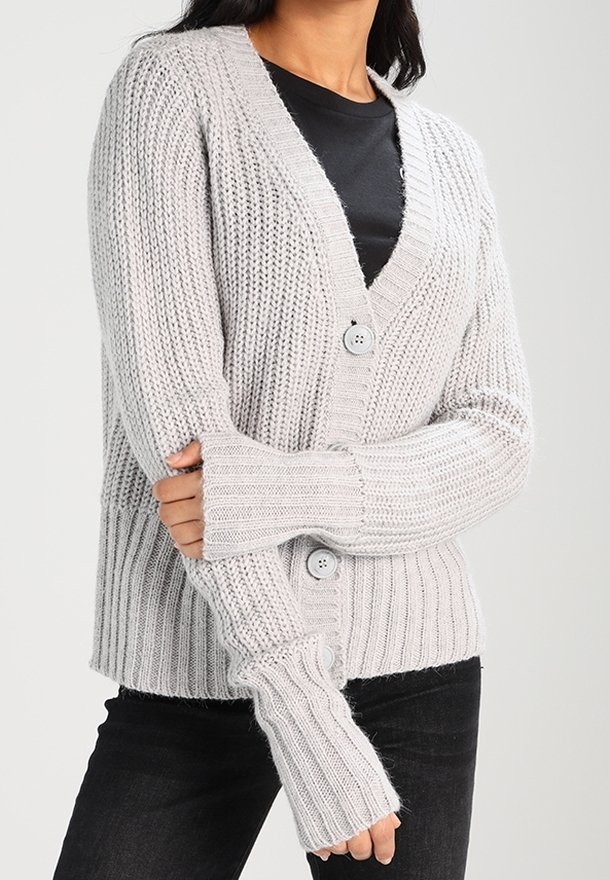 Světlé šedé pletené kardigán s V-výstřihem, ribbovanou texturou, dvěma knoflíky a manžetami s otvory na palce, kombinovaný s černým topem a tmavými džíny.