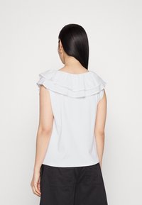 Haut blanc sans manches avec deux rangées de volants au niveau du col et une texture lisse, porté avec un short foncé.