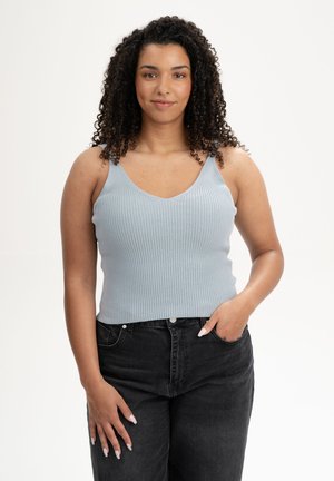 Frau mit lockigem Haar, die ein hellblaues, geripptes Tanktop und schwarze Jeans trägt, steht mit einer Hand in der Tasche vor weißem Hintergrund.