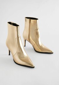 Kultaiset metalliset nilkkasaappaat, joissa on terävä kärki ja stiletto-kanta. Saappaissa on sivuvetoketju ja sileä pinta.
