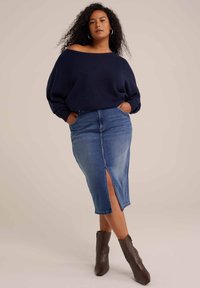 Maglione oversized blu navy con design scollato, abbinato a una gonna tubino in denim con spacco frontale. Stivali alla caviglia marroni.