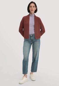 Rostfarbener Strickcardigan mit offener Vorderseite, getragen über einem helllila Rollkragenpullover, kombiniert mit mittelblauen Jeans im geraden Schnitt und weißen Sneakers.