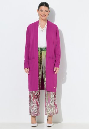 Donna sorridente con cardigan lungo rosa, top bianco, pantaloni ampi con motivo floreale, tacchi bianchi e grandi orecchini rettangolari, in piedi.