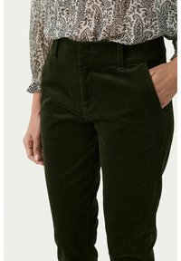 Grüne Cordhosen mit hoher Taille, ausgestattet mit seitlichen Taschen und einem strukturierten Stoff. Kombiniert mit einer gemusterten Bluse.