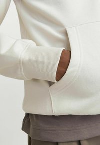 Sweatshirt de couleur crème avec des poignets côtelés et une poche avant, superposé sur une chemise grise foncée. Tissu en coton doux et détails de couture subtils.