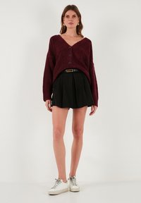 Burgundiszínű textúrájú kardigán V-nyakkal, három gombbal, fekete pliszírozott szoknyával és fehér sportcipőkkel párosítva. Egyszerű és modern stílus.