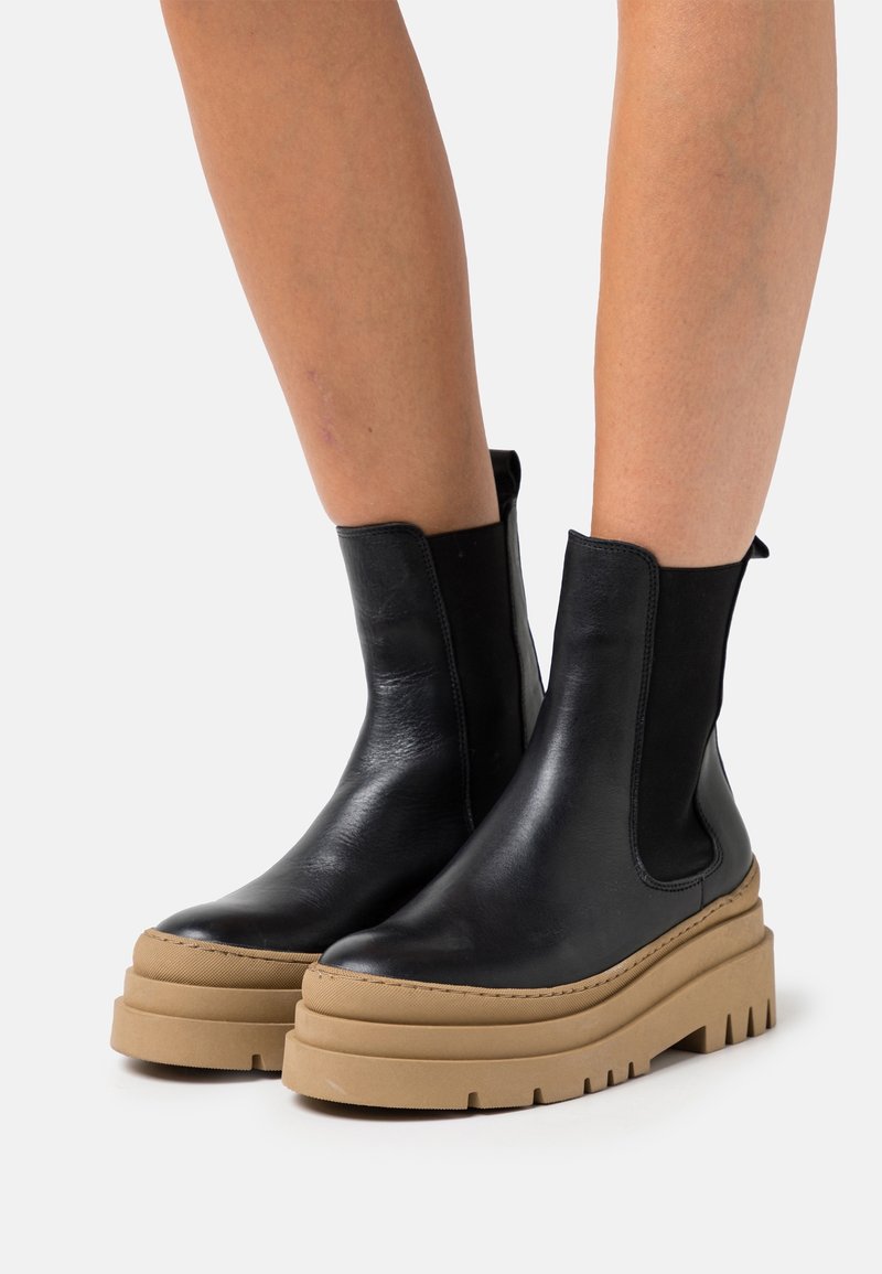 Bottines en cuir noir avec semelles plateformes épaisses couleur camel, portées sur des jambes nues sur un fond gris clair.