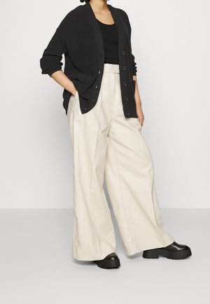 Osoba oblečená v černém kardiganu na knoflíky, černém tričku, širokých béžových kalhotách a černých robustních botách na platformě, stojící na jednoduchém světlém pozadí.