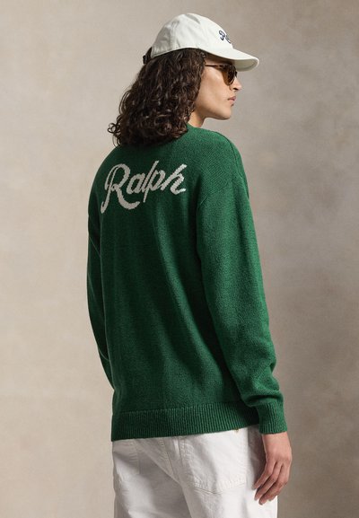 Persona con capelli ricci che indossa un maglione verde con la scritta "Ralph" sulla schiena, pantaloni bianchi, cappello bianco e occhiali da sole, vista di profilo.