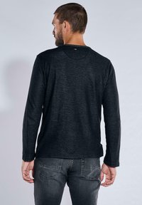Schwarzes Langarmshirt mit rundem Halsausschnitt und gerippter Textur, kombiniert mit dunklen Jeans. Verfügt über ein kleines Logo am hinteren Halsausschnitt.