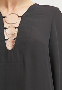 Top nero con scollatura profonda a V, decorato con tre anelli d'argento collegati da fasce nere, realizzato in tessuto liscio con una leggera lucentezza.
