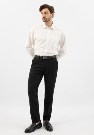 Mann steht mit den Händen hinter dem Rücken, trägt ein weißes Langarmhemd, schwarze Hose, schwarzen Gürtel und schwarze Loafers vor weißem Hintergrund.