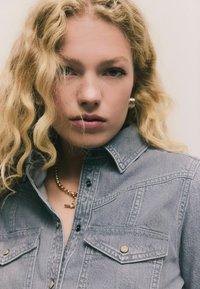 Graues Denim-Hemd mit strukturiertem Kragen, Druckknöpfen und zwei Brusttaschen. Eine goldene Kettenhalskette sorgt für Glanz. Texturiertes Material ist sichtbar.