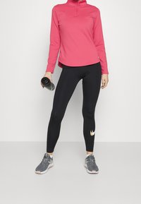 Långärmad, rosa zip-up topp tillsammans med svarta leggings som har en guld månsklogga. Modellen håller en svart vattenflaska och bär grå sneakers.