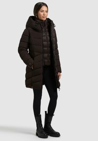 Parka nera con cappuccio, dotata di sezioni trapuntate, chiusura con zip e tasche laterali. Indossata sopra un top nero aderente e leggings, abbinata a stivali neri.