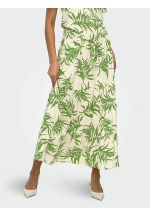Femme portant une jupe midi crème et un top avec imprimé feuilles tropicales vertes, détail noué à la taille, et des talons blancs à bout pointu, debout devant un fond uni.