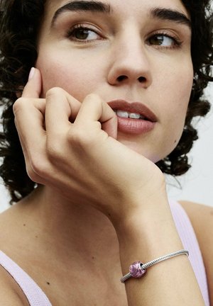 Bracciale d'argento con un ciondolo a forma di rosa rosa. Banda liscia e testurizzata con un accento di fiore dettagliato sulla chiusura. Indossato su un polso contro la pelle.